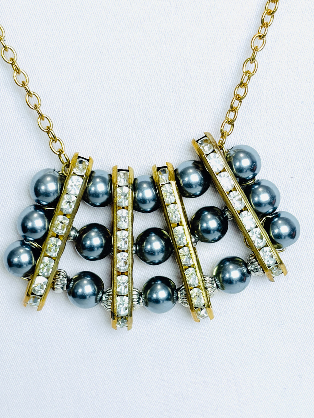 A gold-tone necklace featuring dark grey pearls and clear rhinestone-encrusted bars. Suspended by a simple gold tone rope chain