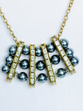 A gold-tone necklace featuring dark grey pearls and clear rhinestone-encrusted bars. Suspended by a simple gold tone rope chain