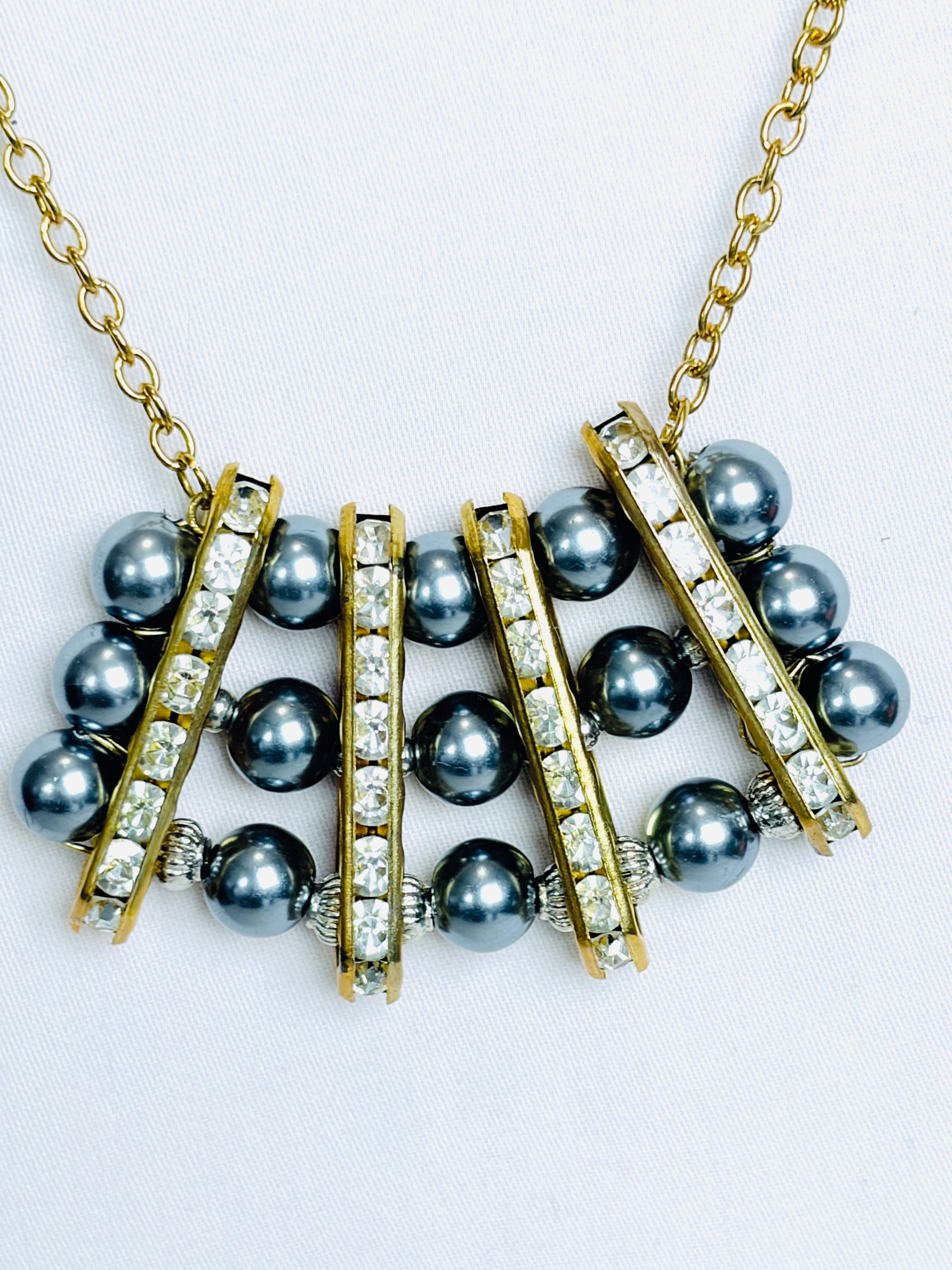 A gold-tone necklace featuring dark grey pearls and clear rhinestone-encrusted bars. Suspended by a simple gold tone rope chain